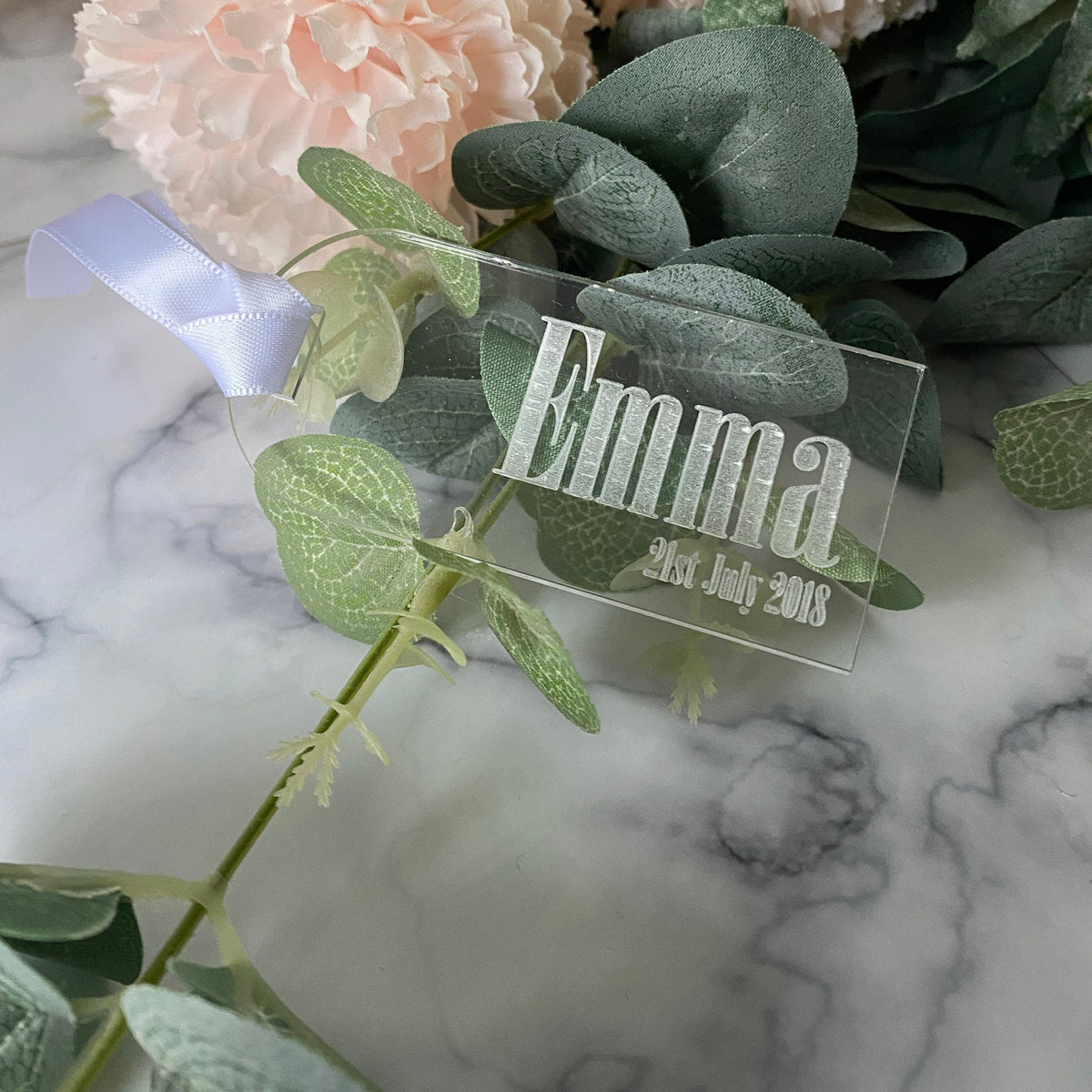 Clear Acrylic Luggage Tag wedding Place Card with 'Emma' and date on a marble surface with flowers and leaves.