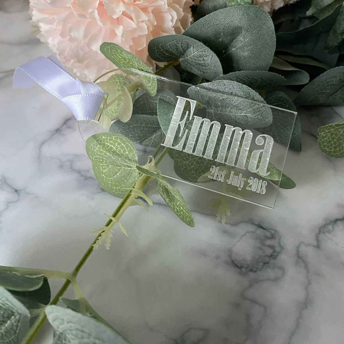 Clear Acrylic Luggage Tag wedding Place Card with 'Emma' and date on a marble surface with greenery