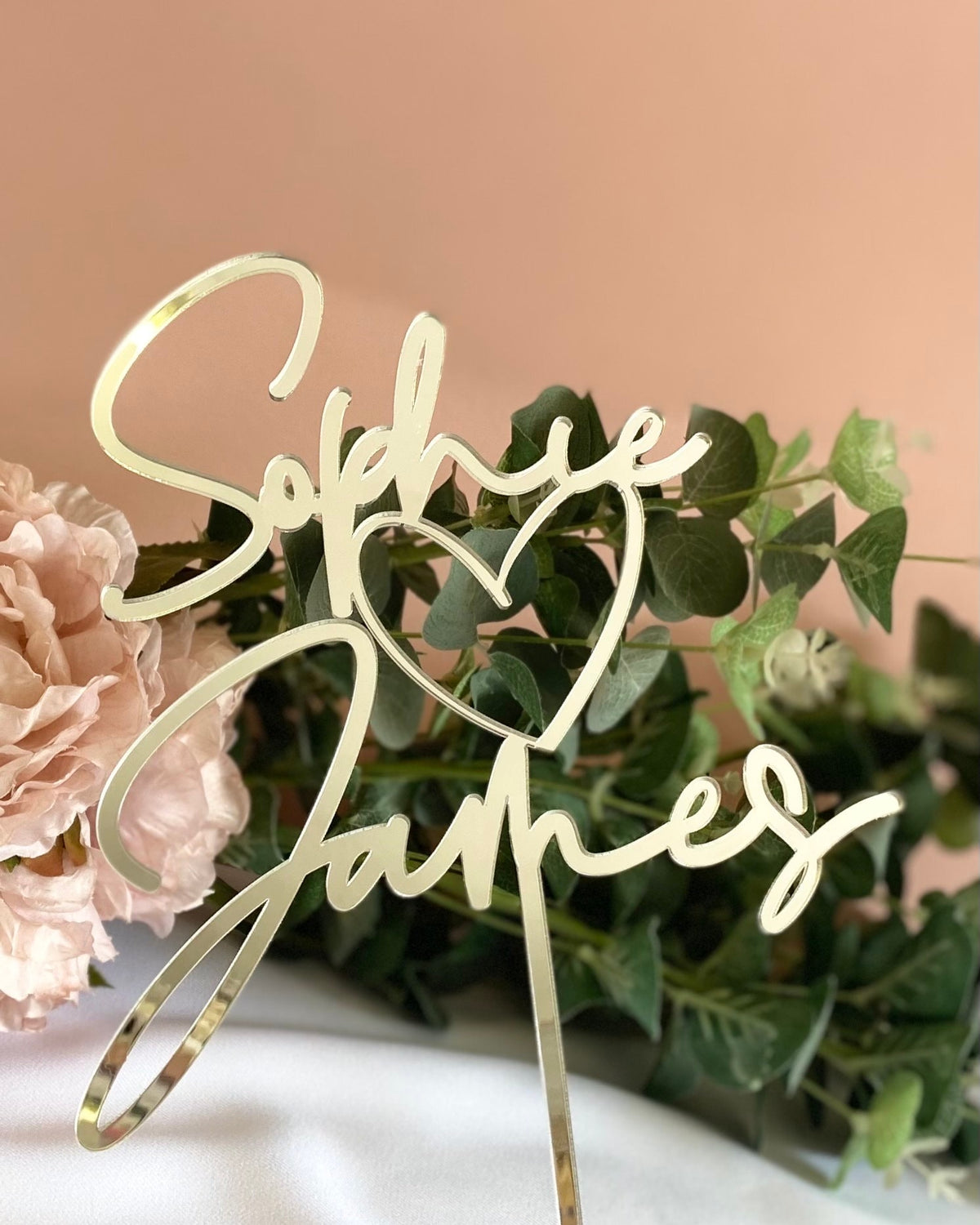 Decorative cake topper with 'Sophie & James' on a pink background with flowers and greenery.