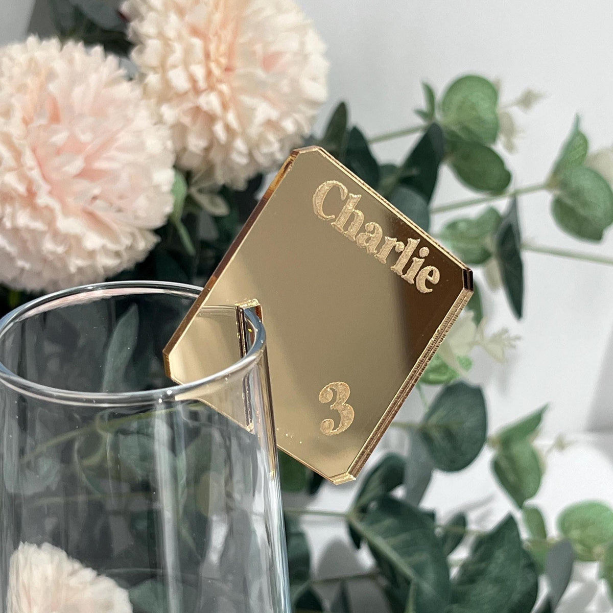 Gold Acrylic Wedding Place Name with 'Charlie' and number '3' on a glass with flowers in the background