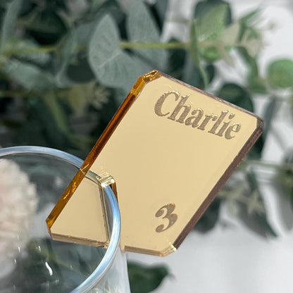Gold mirror Acrylic Wedding Place Name with 'Charlie' and number '3' against a blurred green leafy background