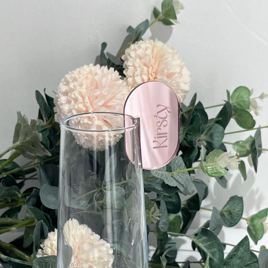 Clear glass rose gold Acrylic Wedding Place Setting - drink marker labeled 'Kirsty' against a background of green leaves and white flowers.