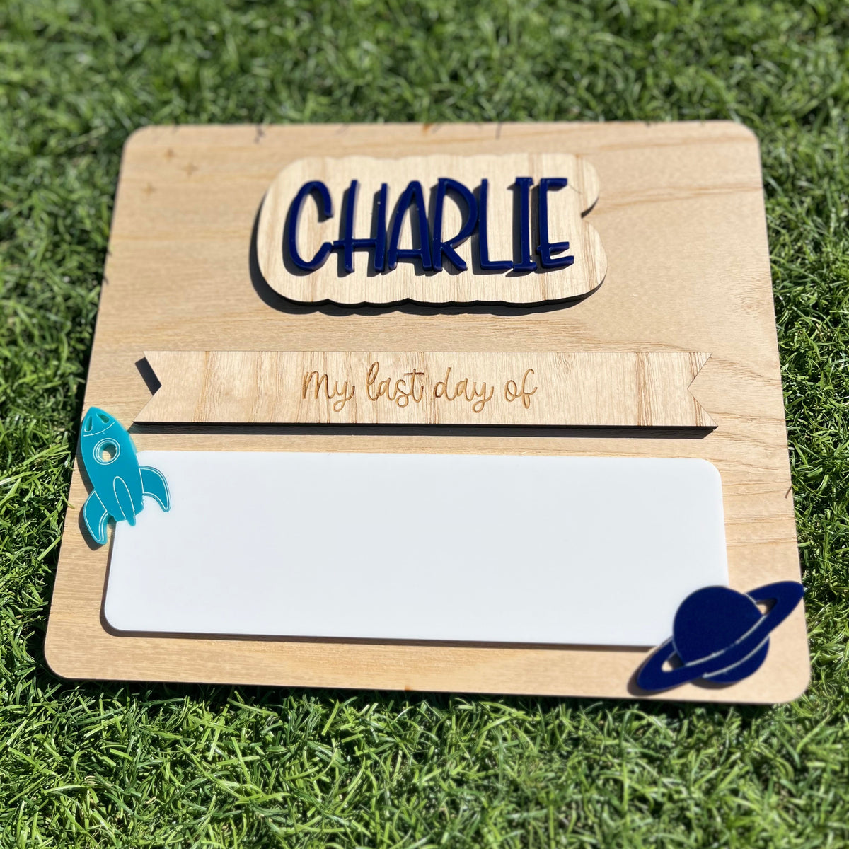 A personalised wooden first/last day plaque featuring interchangeable name plate 'CHARLIE' and 'My last day of', featuring a white reusable writing pad, a rocket ship, and a blue Saturn-like planet.