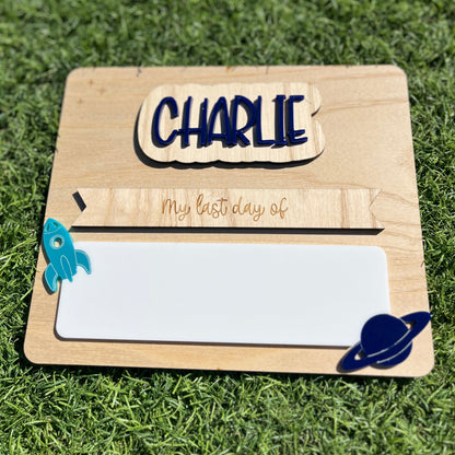A personalised wooden first/last day plaque featuring interchangeable name plate 'CHARLIE' and 'My last day of', featuring a white reusable writing pad, a rocket ship, and a blue Saturn-like planet.