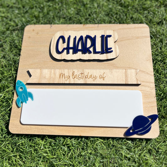 A personalised wooden first/last day plaque featuring interchangeable name plate 'CHARLIE' and 'My last day of', featuring a white reusable writing pad, a rocket ship, and a blue Saturn-like planet.