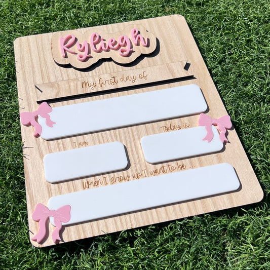 A wooden rectangle shaped personalised plaque with pink decorative bows and white reusable writing spaces 