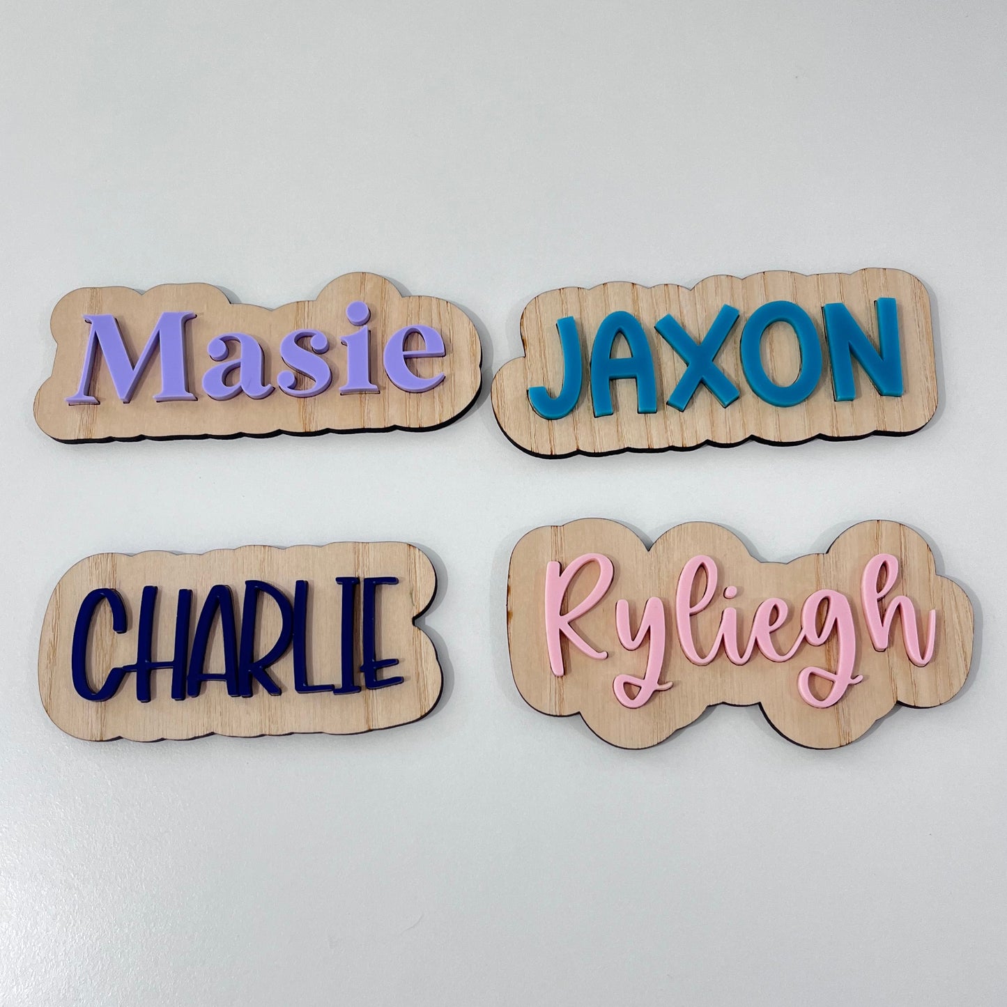 Four personalised wooden nameplates with wavy edges, displayed on a white surface. The names 'JAXON', 'Ryliegh', 'Masie', and 'CHARLIE' are printed in blue, pink, purple, and navy fonts respectively.