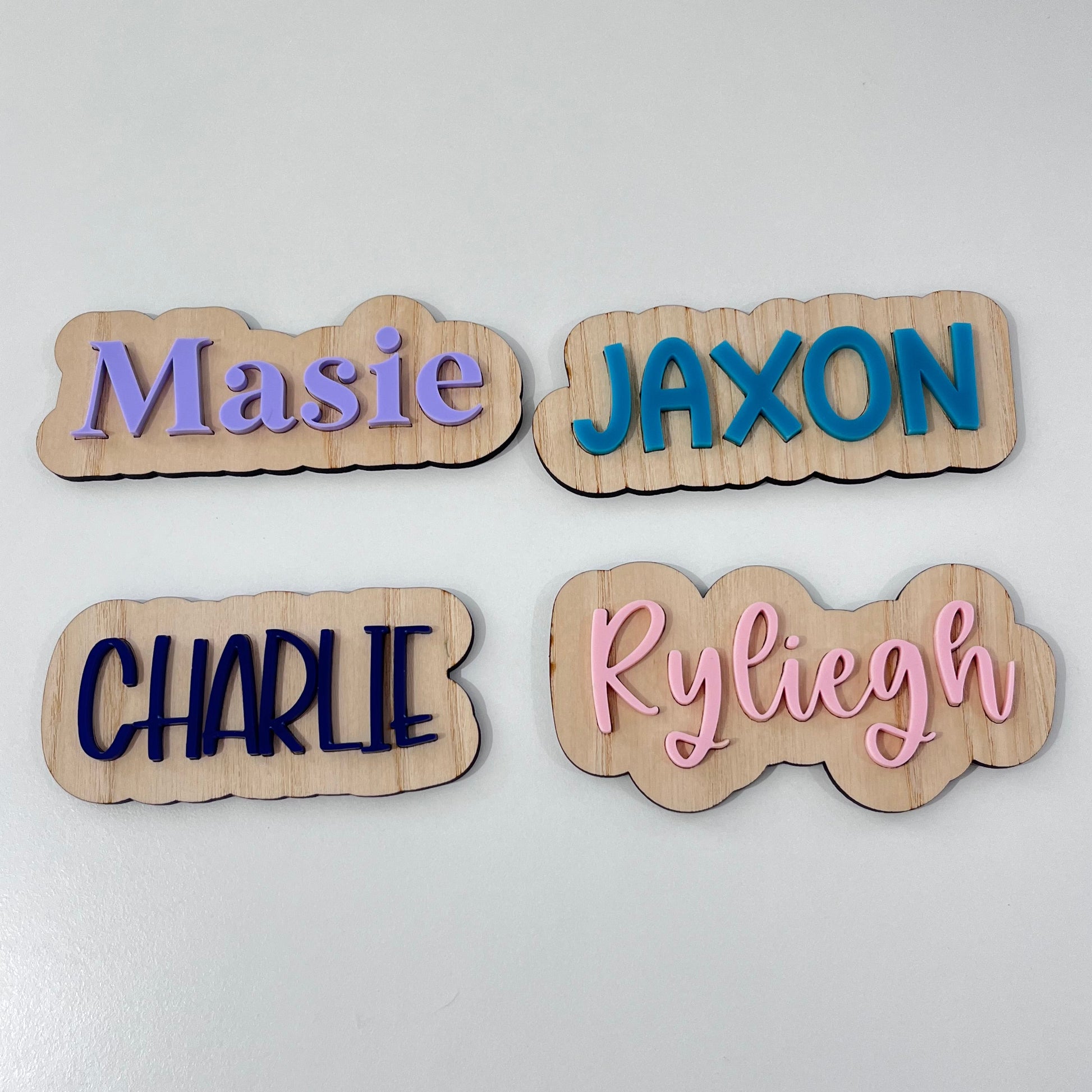 Four personalised wooden nameplates with wavy edges, displayed on a white surface. The names 'JAXON', 'Ryliegh', 'Masie', and 'CHARLIE' are printed in blue, pink, purple, and navy fonts respectively.