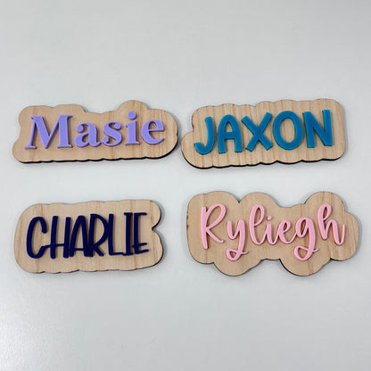 Four personalised wooden nameplates with wavy edges, displayed on a white surface. The names 'JAXON', 'Ryliegh', 'Masie', and 'CHARLIE' are printed in blue, pink, purple, and navy fonts respectively.