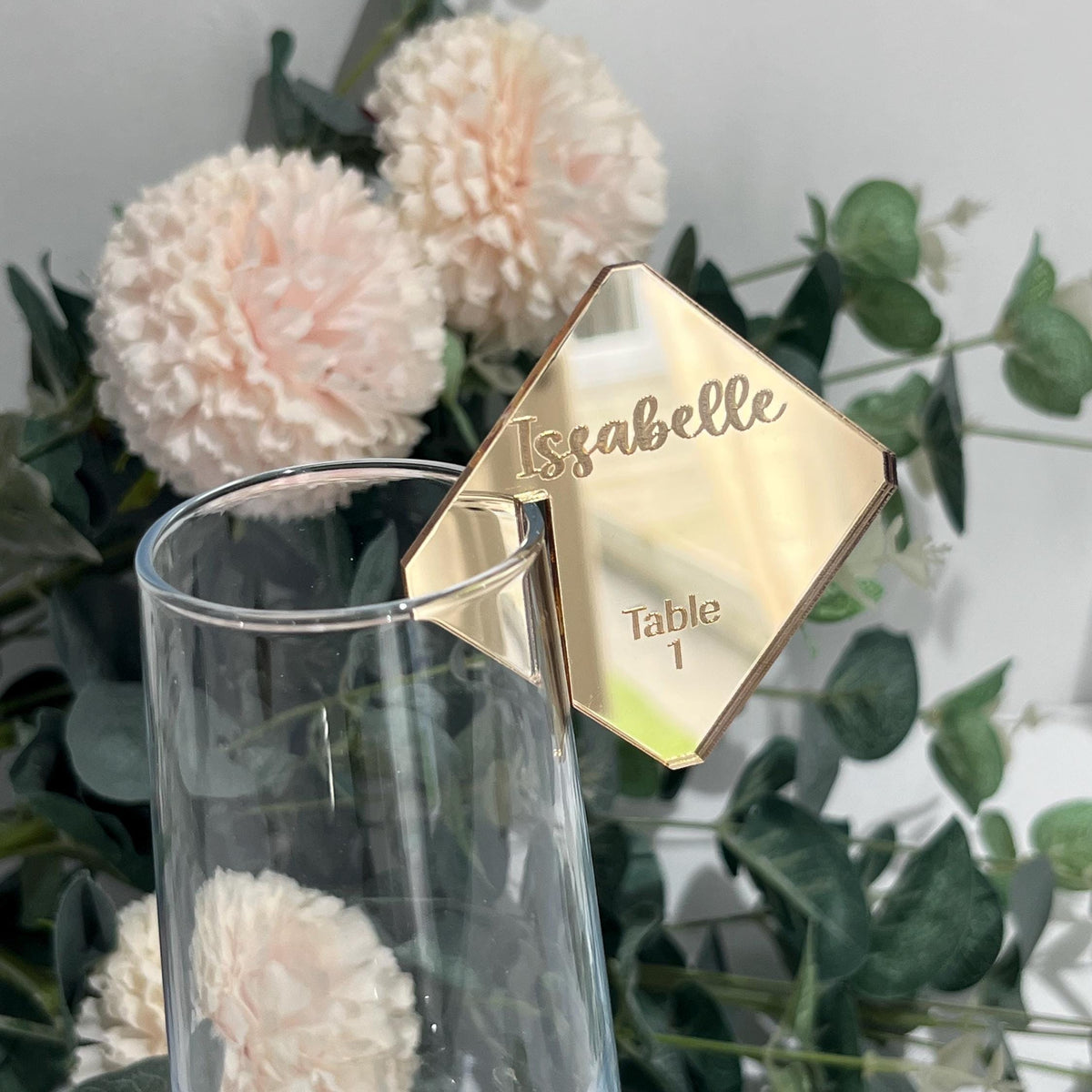 Gold mirror acrylic wedding table name card with 'Isabelle' and 'Table 1' engraved on a floral and greenery background