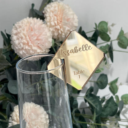 Gold mirror acrylic wedding table name card with 'Isabelle' and 'Table 1' engraved on a floral and greenery background