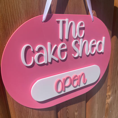 A pink oval hanging sign with white lettering that reads 'The Cake Shed open' attached to a wooden door.