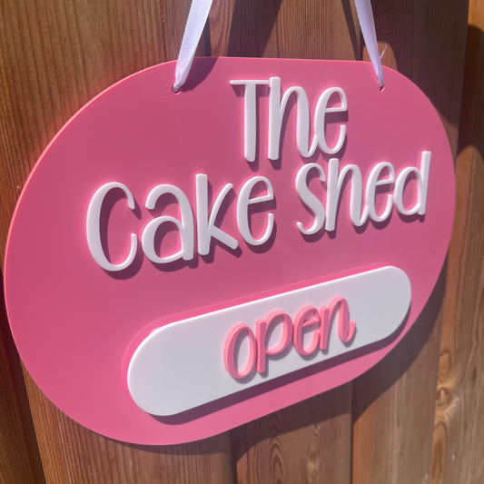 A pink oval hanging sign with white lettering that reads 'The Cake Shed open' attached to a wooden door.
