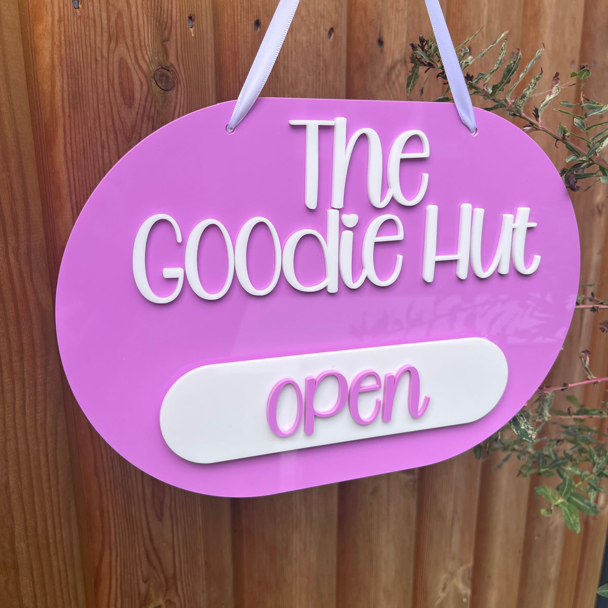 A pink oval hanging sign with white text reading 'The Goodie Hut' and 'open' on a white tab, suspended against a wooden fence.