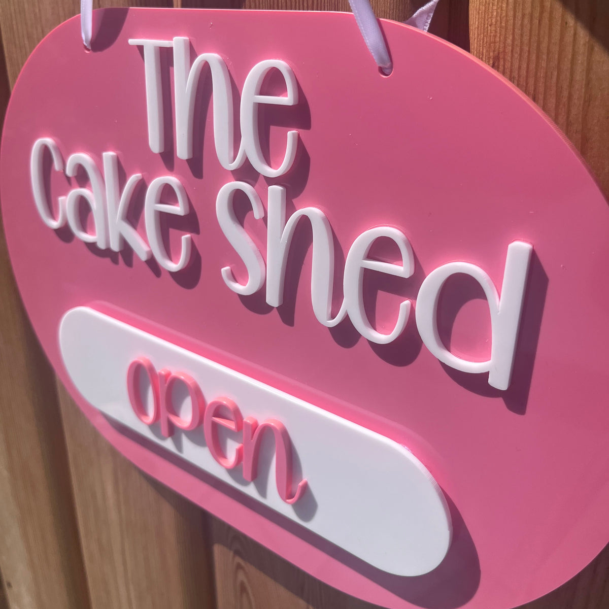 A pink 3d acrylic business sign with white raised lettering that reads 'The cake shed open' against a wooden door background.