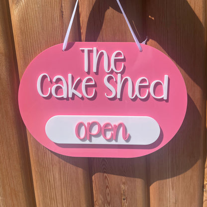 A pink oval acrylic 3d business sign with white text reading 'The Cake Shed open' 
