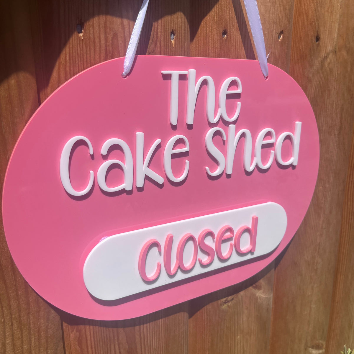 An oval  3d acrylic business hanging sign with white lettering that reads 'The Cake Shed' and 'Closed', mounted against a wooden door.
