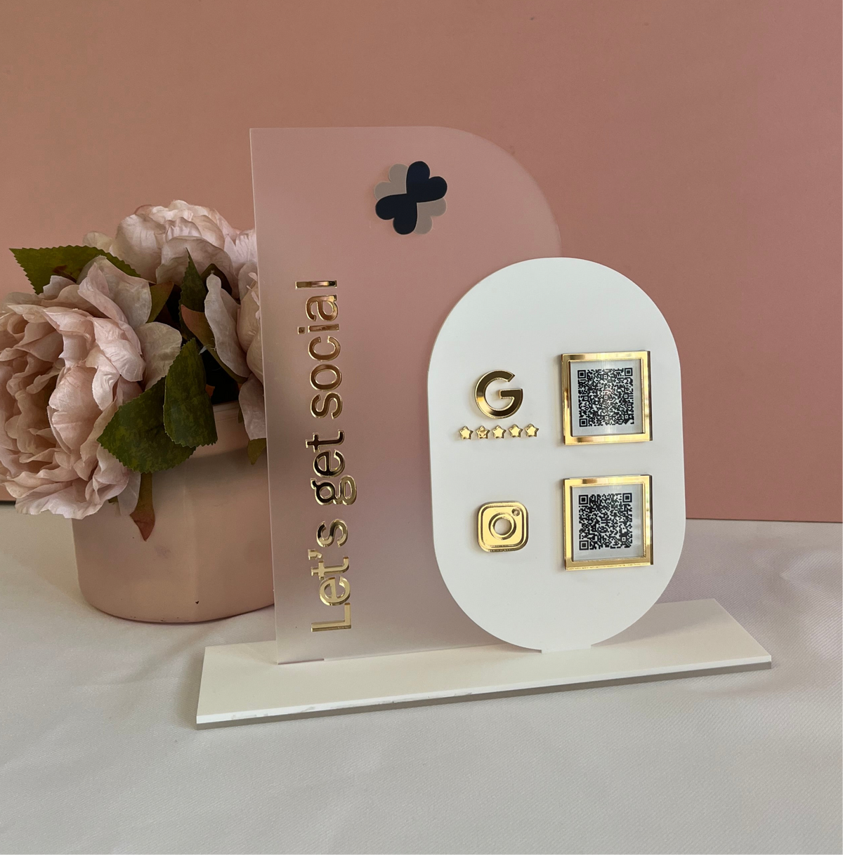 A double layer acrylic QR code sign free standing, with space for a logo and two QR codes, displayed on a white surface against a pink background.