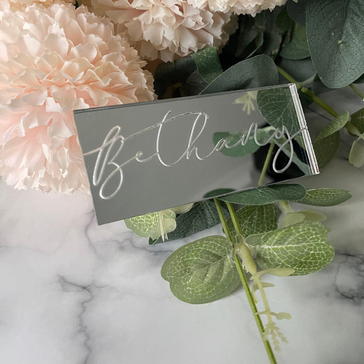 Silver mirror acrylic Engraved Wedding Place Name with 'Bethany' on a marble surface with pink flowers and green leaves.