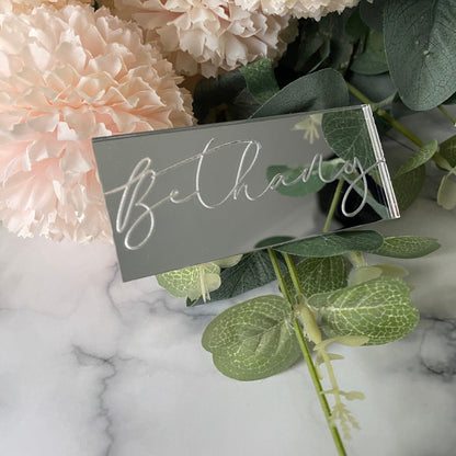 Silver mirror acrylic Engraved Wedding Place Name with 'Bethany' on a marble surface with pink flowers and green leaves.