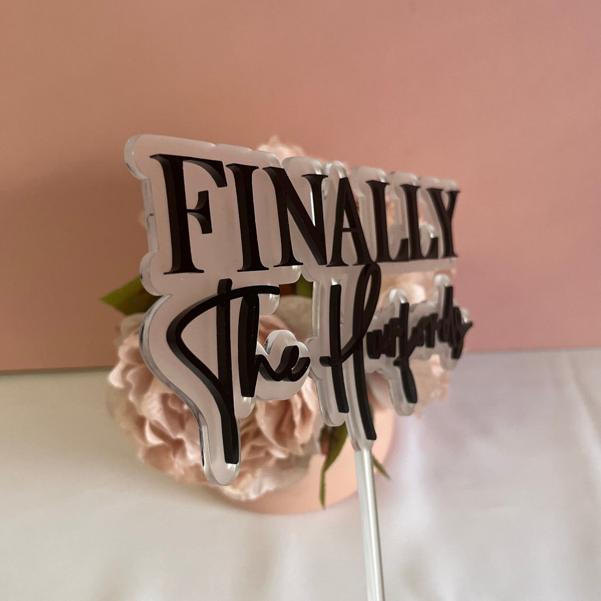An angled view of a Wedding cake topper made with 2 layers of acrylic with the wording Finally the and the couples surname.