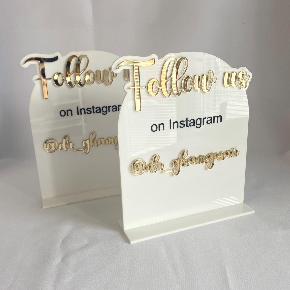 Two acrylic signs with gold text detailing social media handle on a white background