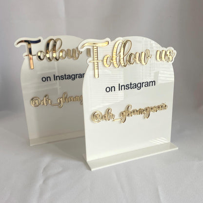 Two acrylic signs with gold text detailing social media handle on a white background