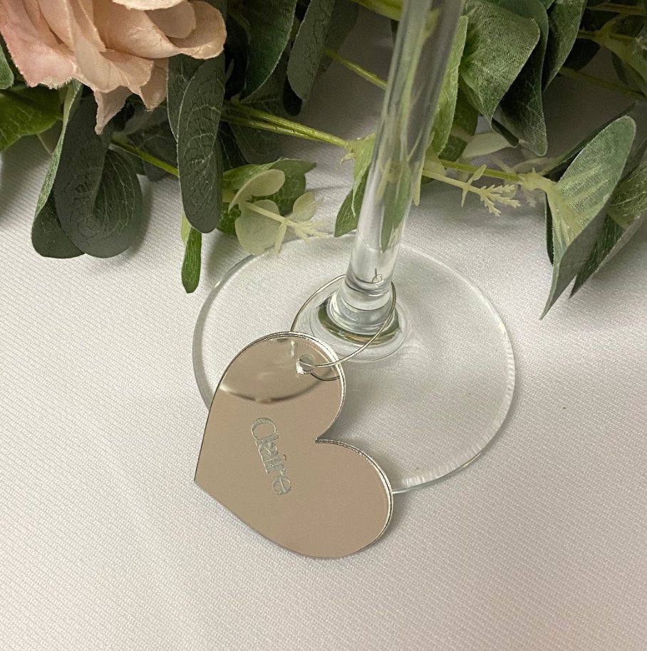 Heart-shaped engraved acrylic wine glass charm on a clear glass with flowers in the background