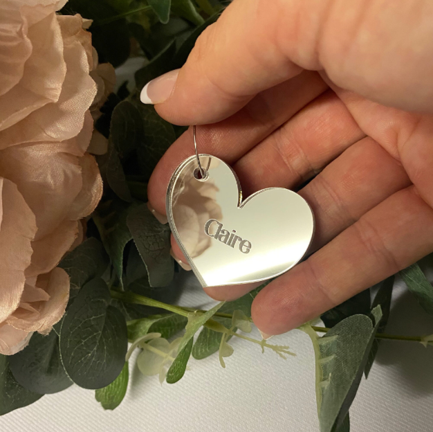 Heart-shaped mirror silver glass charm wedding place setting 'Claire' engraved, held by a hand among flowers.