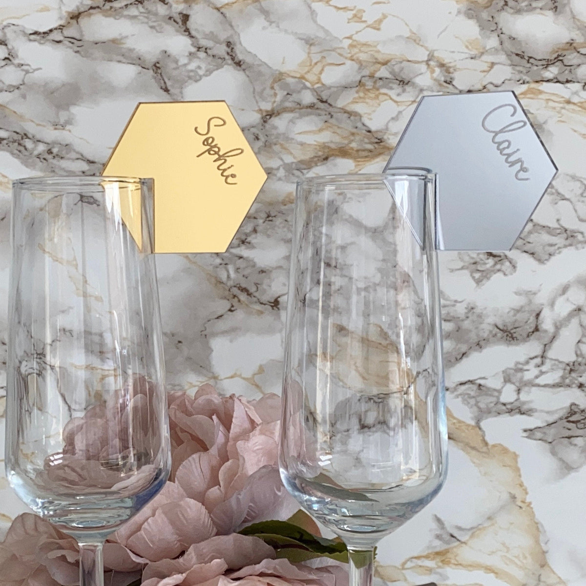 Two clear glass tumblers with gold and silver place cards on a marble surface.
