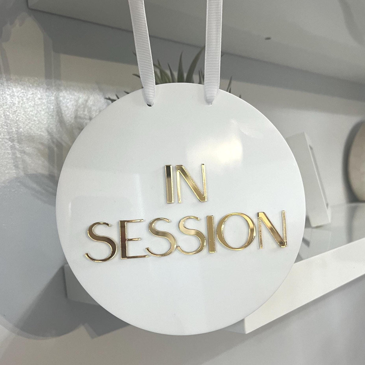 Round acrylic white sign with 'IN SESSION' in gold letters hanging indoors.