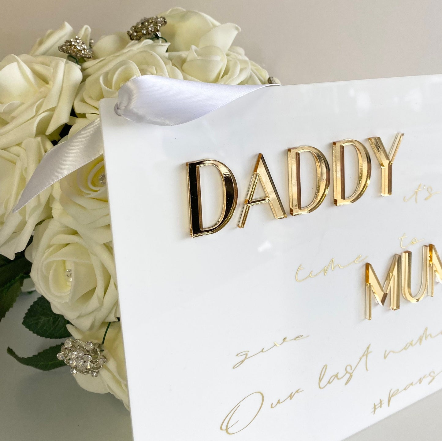 White acrylic sign with gold text 'Daddy it's time to give Mummy our last name text' next to a bouquet of white roses.