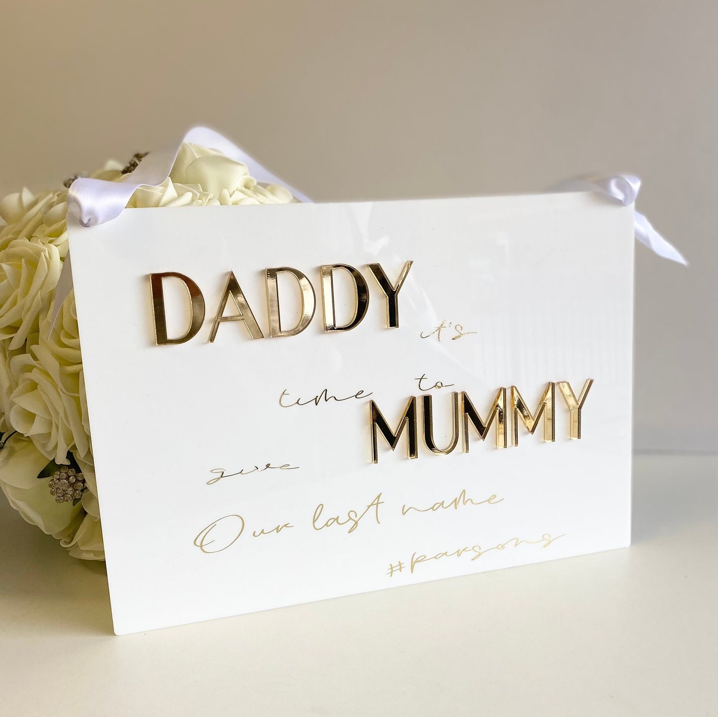 acrylic sign with gold text 'Daddy it's time to Mummy our last name' next to a bouquet of white roses 