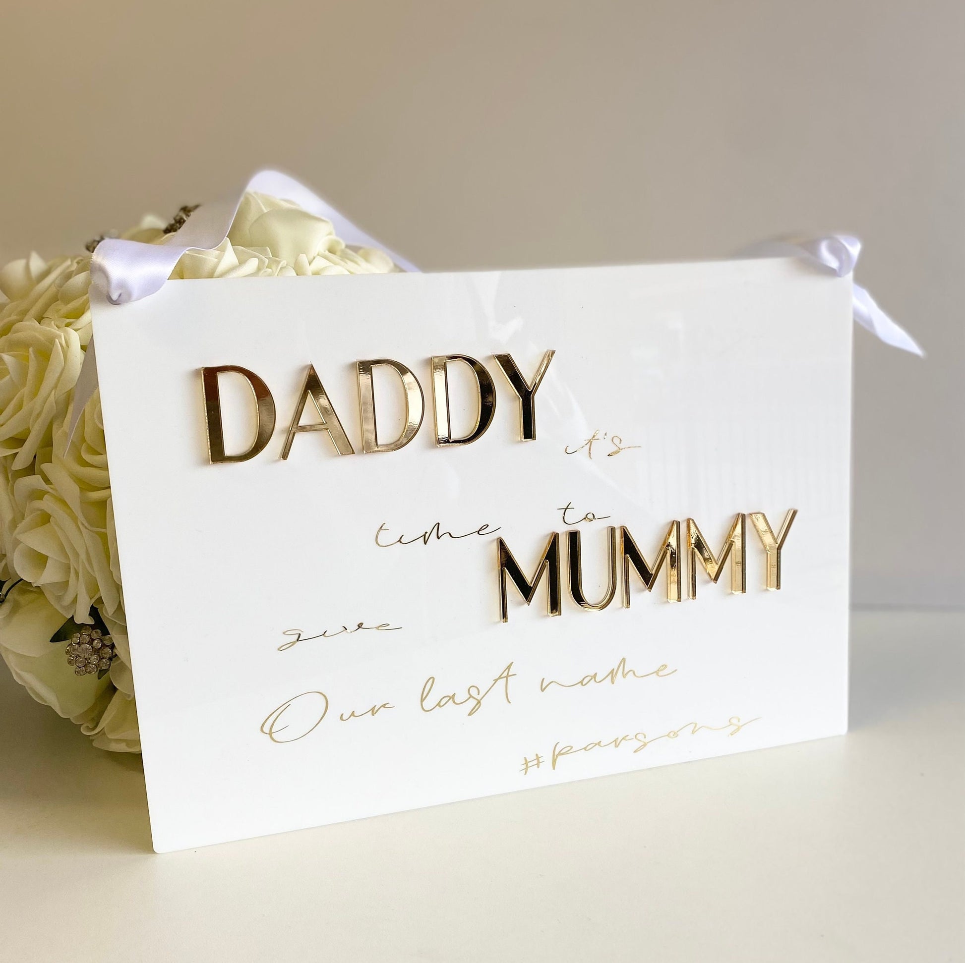 acrylic sign with gold text 'Daddy it's time to Mummy our last name' next to a bouquet of white roses 