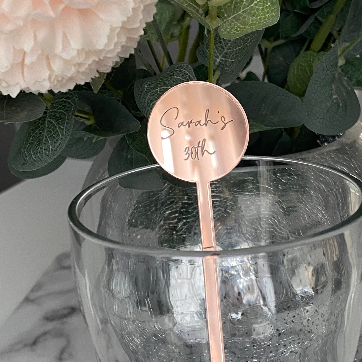 Rose gold cocktail stirrer with 'Sarah's 30th' engraving in a glass with flowers in the background