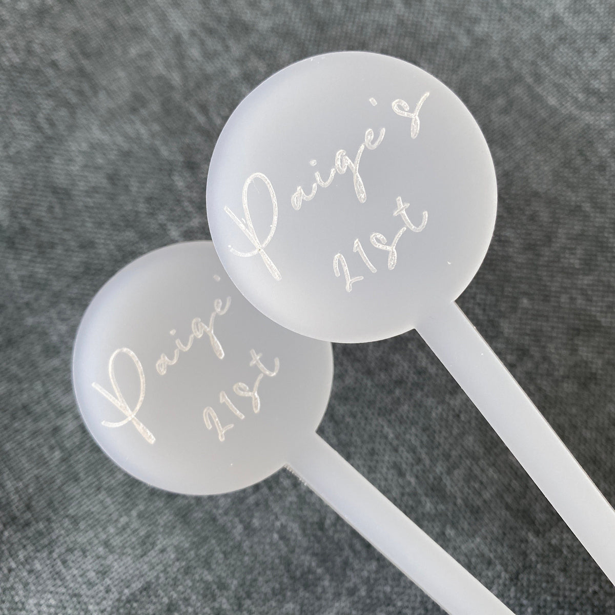 Two white cocktail stirrers with 'Paige's 21st' text on a gray background