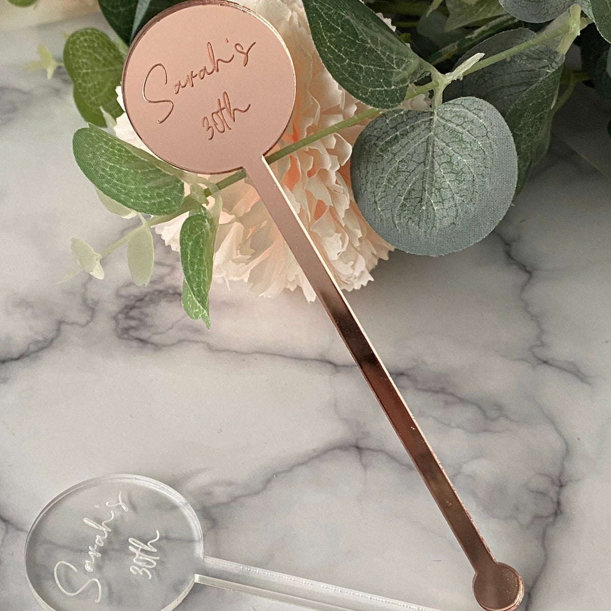 Rose gold and clear drink stirrers with 'Sarah's 30th' engraving on a marble surface with greenery.