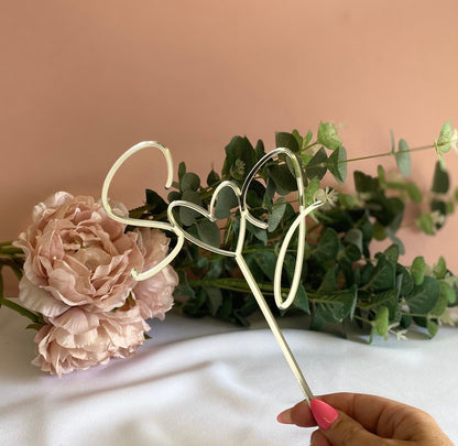 silver acrylic heart and initials cake topper held by a hand against a backdrop of pink flowers and greenery.