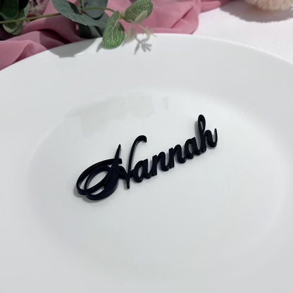 Black acrylic Personalised Laser Cut Wedding Place Name' Hannah' on a white wedding dinner plate with pink fabric and green leaves in the background