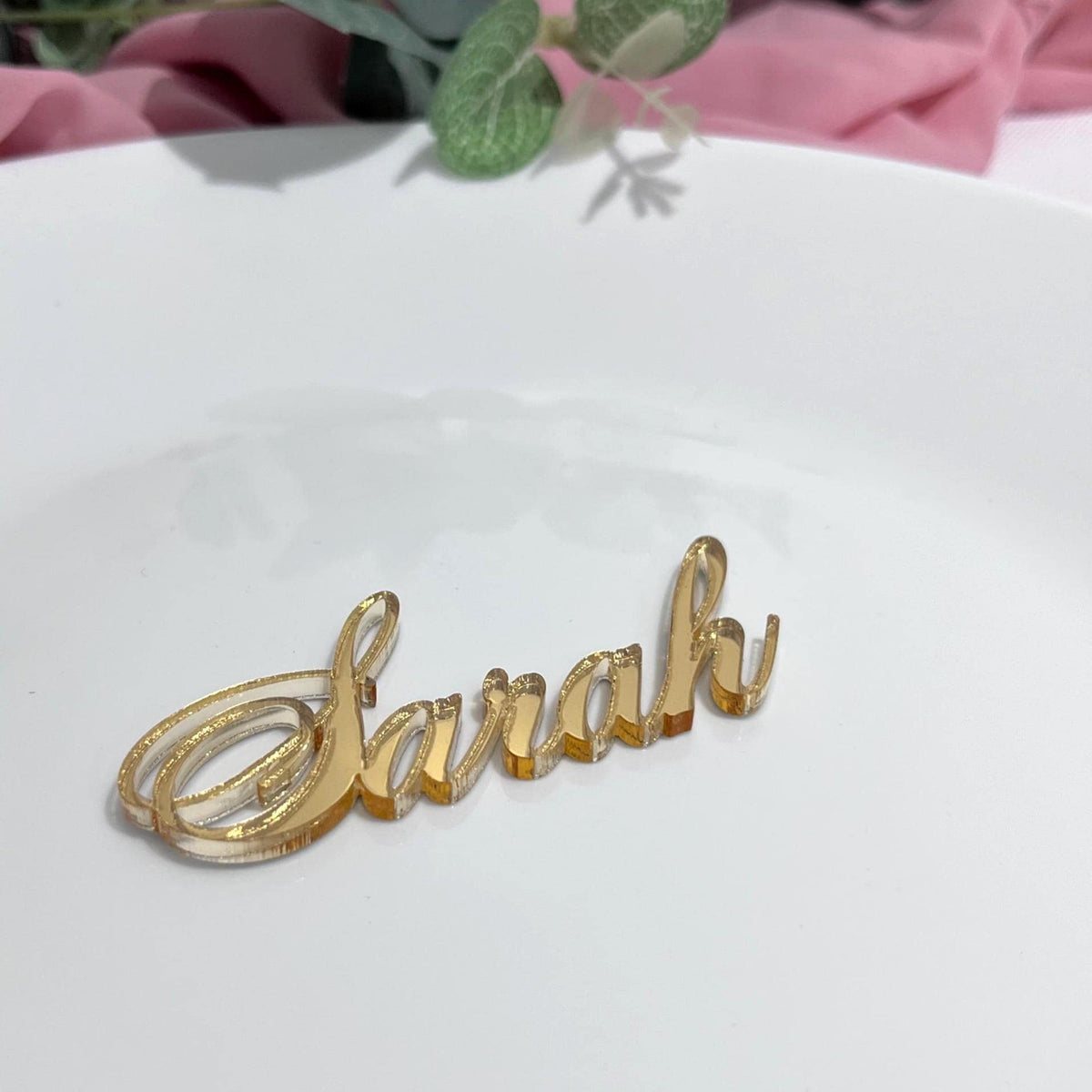 Gold mirror acrylicPersonalised Laser Cut Wedding Place Name 'Sarah' on a white wedding dinner plate with pink fabric and green leaves in the background