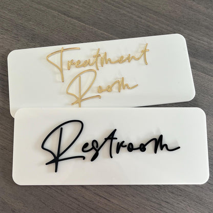 Two business door signs on a wooden surface, one labeled 'Treatment Room' and the other 'Restroom'. Both signs are made from acrylic and have 3d text