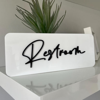 Rectangular white business sign with 'Restroom' text on a shelf with a plant in the background
