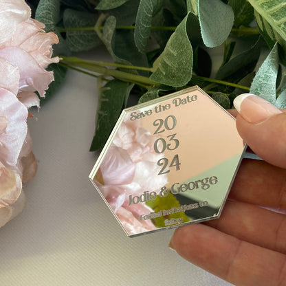 Hexagonal wedding save the date card held by a hand with greenery and flowers in the background.
The save the date is made from mirror silver acrylic with the details engraved.