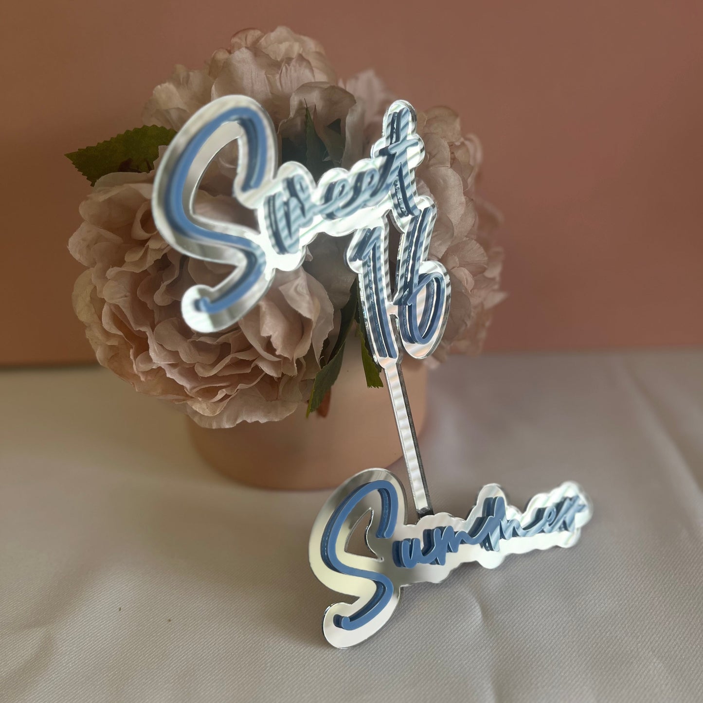 A double layer acrylic cake topper with the words 'Sweet 16' in bold script, placed on a surface with a floral decoration in the background.