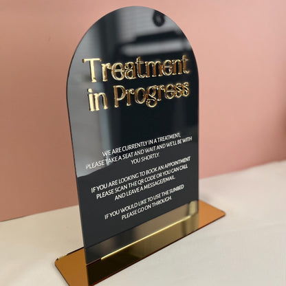 Sign indicating 'Treatment in Progress' with gold text on a black background, placed on a stand against a pink wall.