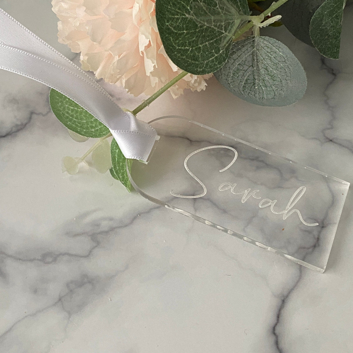 Clear acrylic Wedding Place Name with 'Sarah' engraved, ribbon, and flowers on marble surface