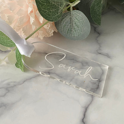 Clear acrylic wedding name tag with 'Sarah' engraved on a marble surface with flowers.