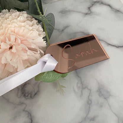 Rose gold acrylic Wedding Place Name with 'Sarah' engraved, ribbon, and flowers on a marble surface