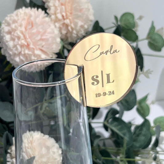 Gold  mirror acrylic round Wedding Place Setting Glass Charms with 'Carla S/L' and '19-9-24' on a clear glass with flowers in the background