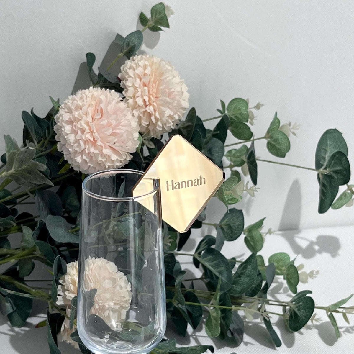 Clear glass with a gold Wedding seating chart Name tag labeled 'Hannah' surrounded by flowers and greenery on a light background